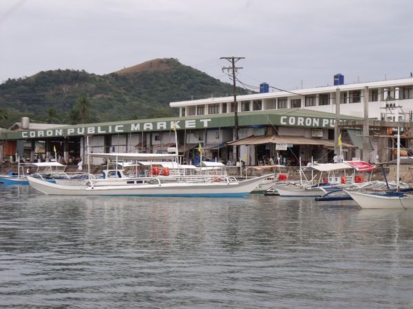 Long green-roofed market building with Coron Public Market painted across the facade, outrigger boats moored in front