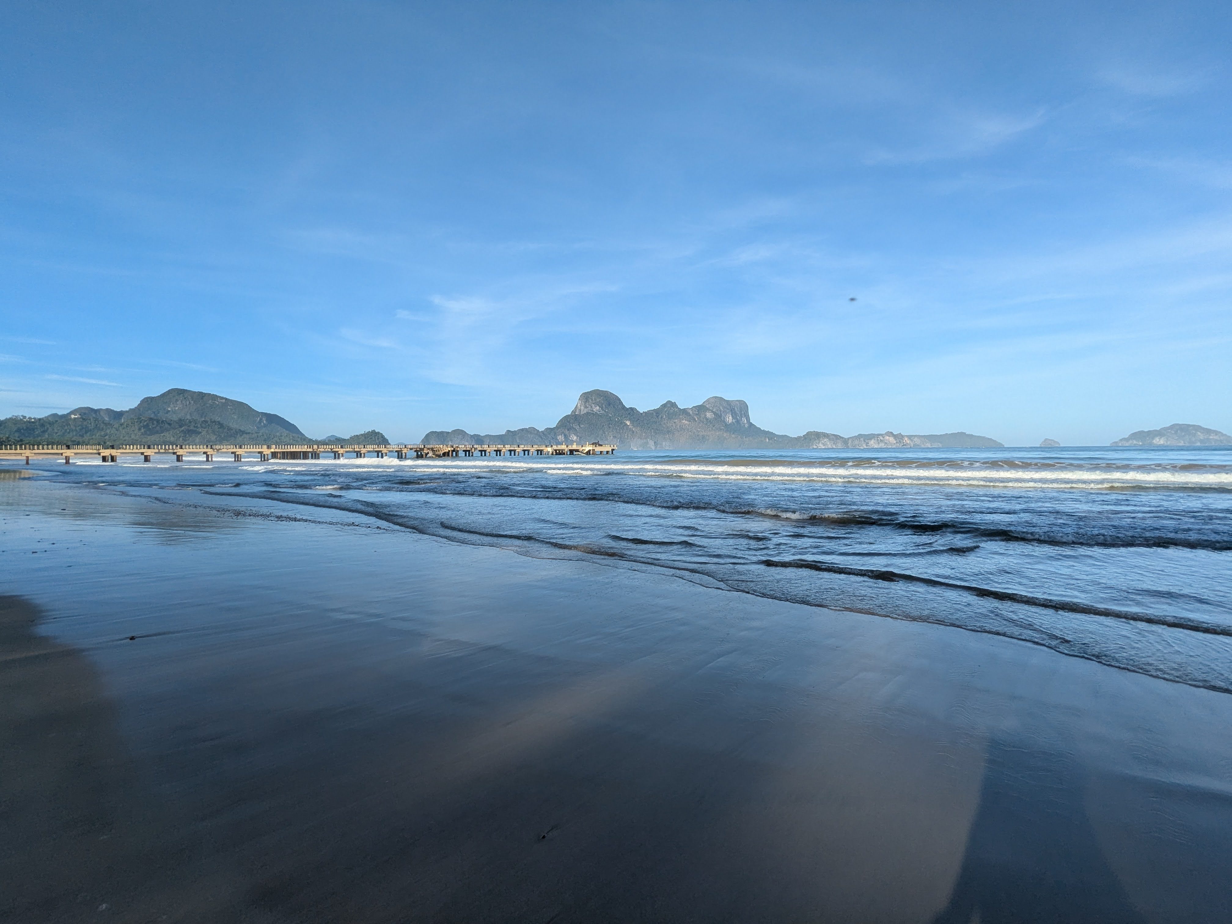 Low tide at Lio Beach with wet flat sand, small breaking waves, the long pier on stilts and Cadlao Island on the horizon