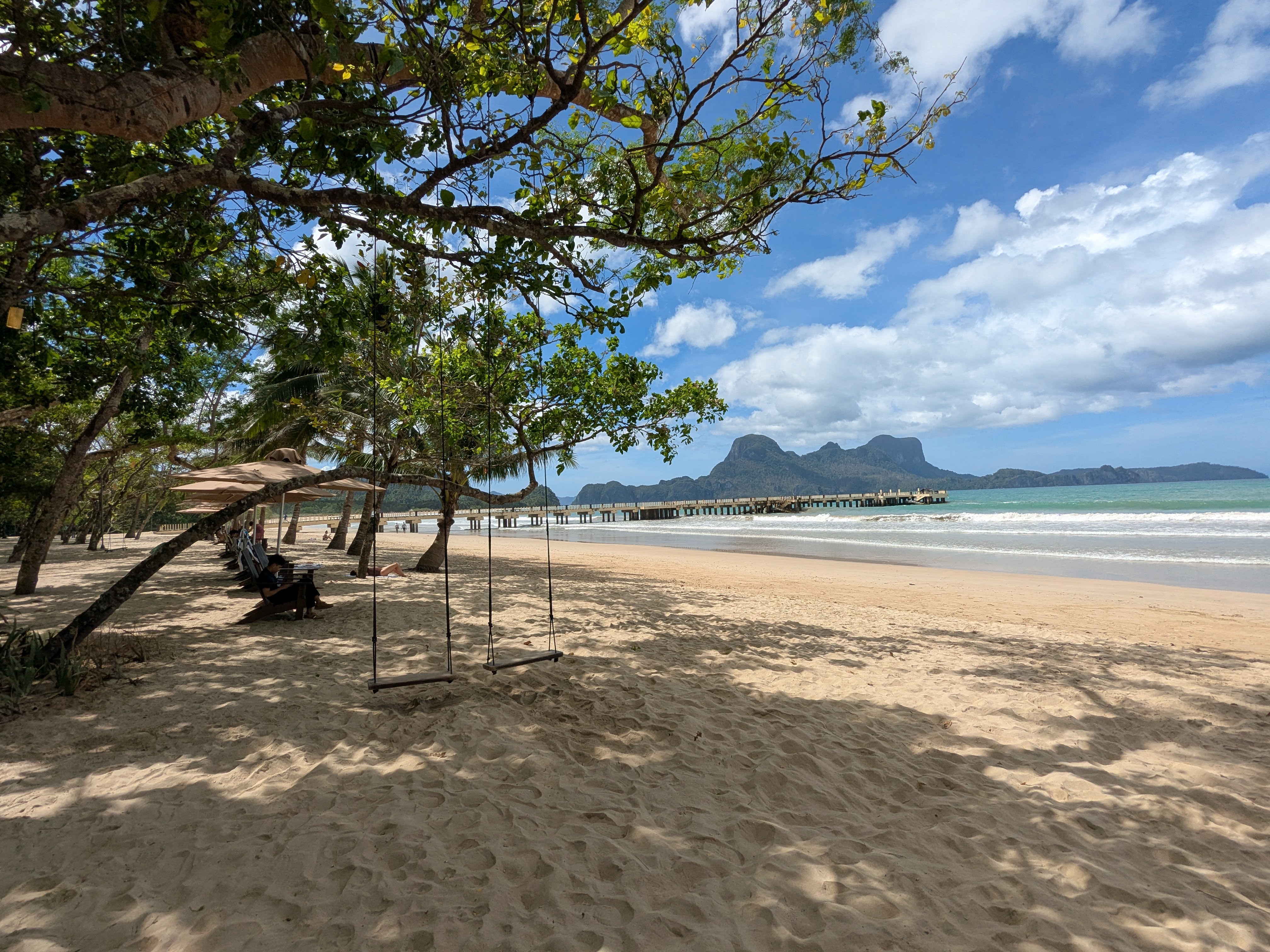 White sand at Lio Beach with a wooden swing hanging from a shore tree, loungers under umbrellas and the long pier reaching into the bay