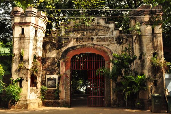The ruined masonry portal of Plaza Cuartel with flanking towers, exposed brickwork and a red iron gate standing open