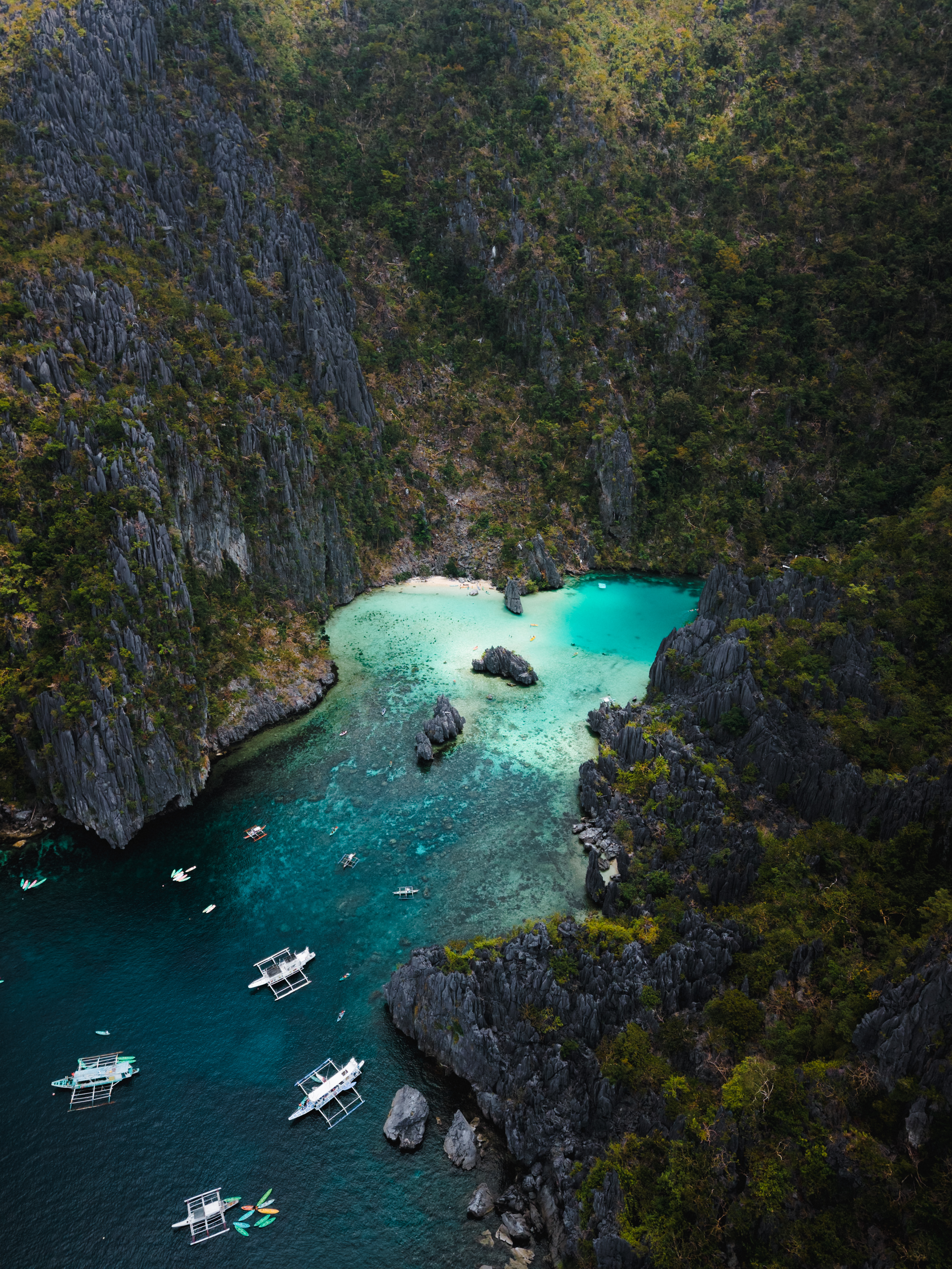 Aerial view of Cadlao Lagoon, a turquoise inlet reached through a narrow gap between limestone cliffs, with outrigger boats moored outside
