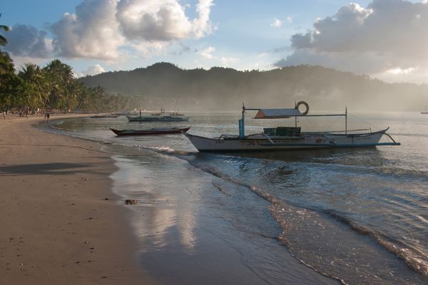 Outrigger bangka boats moored at the waterline on the sandy shore of Port Barton bay in late afternoon light, with a palm-lined shore and forested hills behind