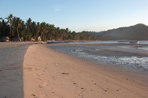 Wide sandy beach at Port Barton at golden hour, with a rippled tidal flat, outrigger boats drawn up along the shore and coconut palms and village huts along the treeline