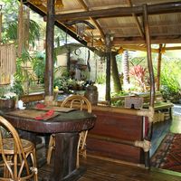 Open-air bamboo and hardwood dining area with rattan chairs around a dark wooden slab table, bamboo wind chimes and a tropical garden beyond