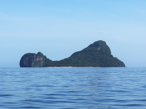 Helicopter Island in Bacuit Bay seen from the water, a low forested island with a rocky hump at one end and a white sand strip along its base