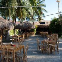 Outdoor courtyard with wooden tables and rattan chairs under nipa thatched huts, palm trees and star lanterns strung from a bamboo pole