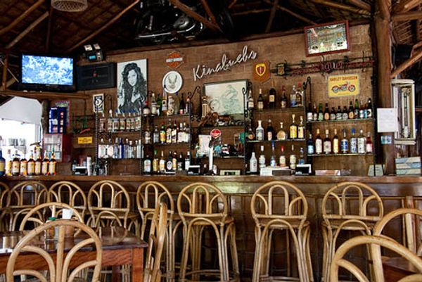 The back bar with the Kinabuchs name in white script across the wooden wall, spirit bottles on shelves and empty rattan bar stools
