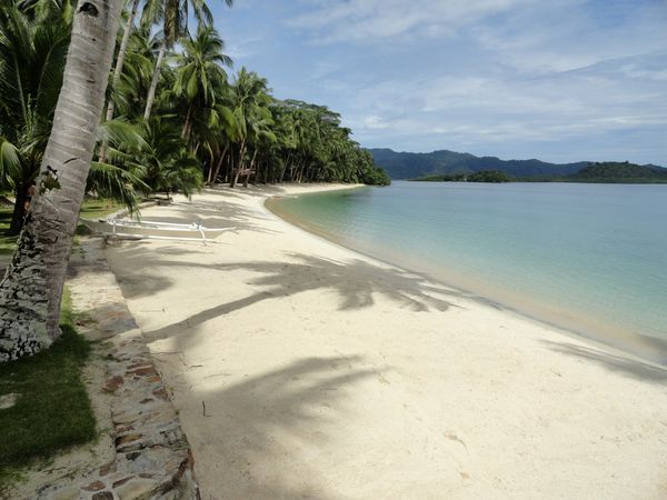 A curving white sand beach backed by dense coconut palms, with two white outrigger boats pulled up on the sand and forested hills across the water